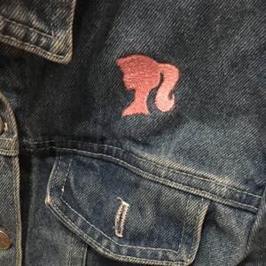 May include: A blue denim jacket with a pink Barbie logo embroidered on the front pocket.