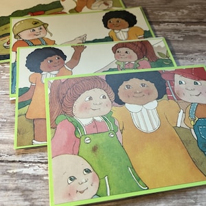 May include: A set of four rectangular cards featuring colorful illustrations of children with various hairstyles and clothing, including overalls and dresses, with a light green border around each card, stacked on a weathered wooden surface.