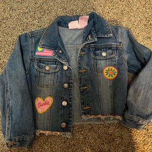 May include: A blue denim jacket with patches. The patches are a pink heart with the word "Barbie" inside, a yellow, blue, and red flag, and a yellow flower with a green center.