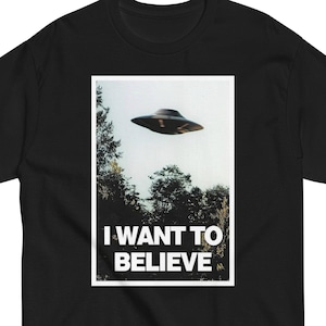 May include: Black t-shirt featuring a rectangular graphic with a flying saucer over a forest. The graphic includes the text "I WANT TO BELIEVE" in white block letters. The shirt is made of a soft material.