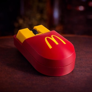 Peut inclure: Une souris d'ordinateur rouge en forme de boîte de frites McDonald's, avec des boutons jaunes ressemblant à des frites. Le logo McDonald's est affiché en jaune. La souris est sur une surface marron foncé.