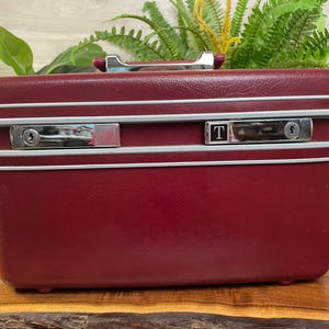 May include: A rectangular burgundy train case with a textured finish, featuring a silver-toned top handle and two metallic locking latches on the front. One latch displays a square black emblem with the white letter T. The case has light gray trim.