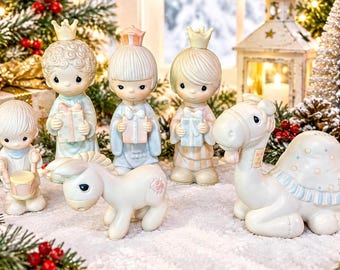 Może przedstawiać: Zestaw porcelanowych figurek przedstawiający troje dzieci w koronach trzymających pudełka z prezentami, małe dziecko grające na bębnie, osiołka i wielbłąda. Figurki są pomalowane w delikatnych pastelowych odcieniach błękitu, różu i kremu.