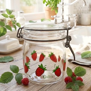 May include: A clear glass storage jar with a metal wire bail and lever closure. The jar features a repeating pattern of red raspberries with green leaves printed on the exterior. The text The Cannery is printed in green near the bottom base.