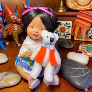 May include: A handcrafted doll with black hair and a pink bow, wearing a white top and a colorful skirt. The doll holds a small white teddy bear with an orange vest and blue bow tie. Decorative items are in the background.