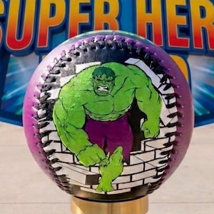 Puede incluir: Una pelota de béisbol con un gráfico de Hulk verde sobre un fondo morado y negro. Hulk está representado rompiendo una pared de ladrillos. La pelota tiene costuras blancas y una base dorada.