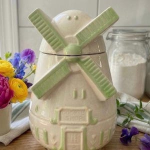 Könnte beinhalten: Ein Keramik-Keksdose in Form einer Windmühle mit cremefarbenem Körper und hellgrünen Akzenten an den Flügeln, der Tür und den Fensterrahmen. Auf der Vorderseite befindet sich ein erhabenes Schild mit dem geprägten Text COOKIE SHOP.