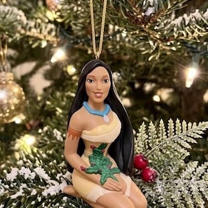 May include: A Christmas tree ornament featuring a Native American woman in a kneeling pose. The ornament has long black hair, a tan complexion, and is wearing a cream-colored dress with a blue necklace. A green leaf design is on the front.