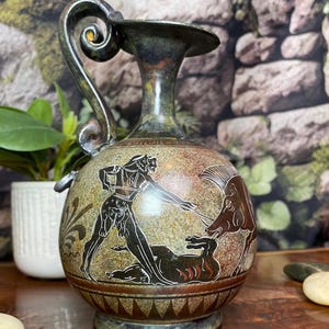 May include: A decorative ceramic pitcher featuring a textured, mottled green and brown finish. The body displays a black-figure style illustration of a figure in combat with a wild boar. It has a curved handle and a wide, flared rim.