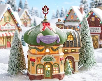 Może przedstawiać: Miniaturowy budynek North Pole Beauty Shoppe z zielonym kopułowym dachem, czerwoną kokardą i dzwonkiem na szczycie. Konstrukcja posiada żółte ściany, ceglane akcenty, podświetlane okna i fioletowy baner z białym napisem. Dwie ośnieżone sosny stoją po obu stronach budynku w zimowej scenerii.