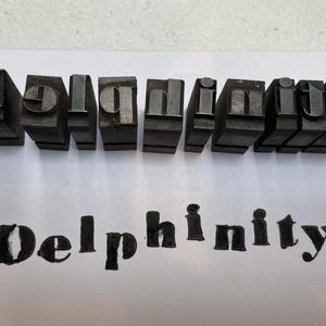 May include: A set of ten individual metal letterpress blocks arranged in a horizontal row, spelling the word Delphinity in reverse. Below the metal blocks, the word Delphinity is printed in black ink on a white surface, showing the final result.