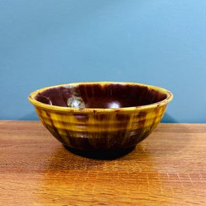 Puede incluir: Un cuenco de cerámica con un patrón de rayas marrones y amarillas. El cuenco tiene un interior marrón oscuro y una forma redondeada. El cuenco está sobre una superficie de madera.