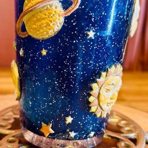 May include: A blue glass tumbler with a celestial design. The tumbler features a starry night sky with gold planets, stars, and a sun with a face. The glass is set on a decorative metal coaster.