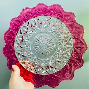 May include: A stack of decorative glass plates. The bottom plate is a vibrant magenta with a scalloped edge. The middle plate is clear with a starburst pattern. The top plate is a clear, ruby red.