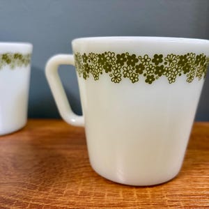 Puede incluir: Taza de cerámica blanca con asa, adornada con un borde floral verde oliva cerca del borde. La taza está sobre una superficie de madera, con otra taza visible en el fondo.