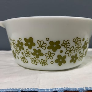 May include: White Pyrex bowl with an olive green floral pattern. The bowl has two handles and a rounded shape. The floral design is a repeating pattern of stylized flowers and leaves around the bowl's exterior.