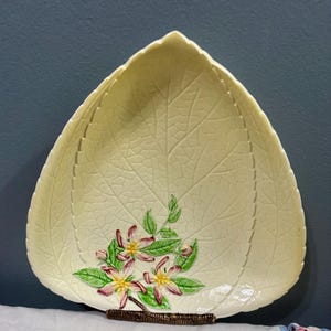 Puede incluir: Plato de cerámica con forma de hoja en un tono amarillo pálido, con un patrón de venas detallado. El plato está adornado con un grupo de flores rosas y amarillas con hojas verdes. Se apoya en un soporte decorativo marrón.