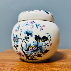 以下が含まれることがあります： クリーム色の蓋付きセラミックジャー。花柄で飾られています。 デザインは、黄色の中心、ピンクのアクセント、緑の葉を持つ大きな青い花を特徴としています。 ジャーは木製の表面にあります。