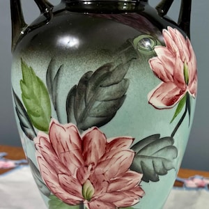 Puede incluir: Un jarrón de cerámica con fondo azul claro y dos asas. El jarrón está decorado con flores rosas y hojas verdes. La parte superior y las asas son de color marrón oscuro.