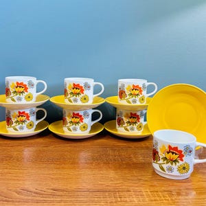 Könnte beinhalten: Ein Set aus sechs weißen Keramik-Teetassen und Untertassen mit einem floralen Design in Rot, Gelb und Orange. Die Untertassen sind leuchtend gelb. Eine Tasse und Untertasse sind separat.
