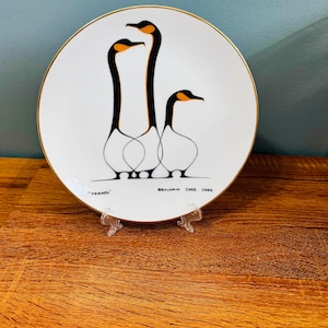 Puede incluir: Plato decorativo blanco con borde dorado. Presenta una ilustración minimalista de tres gansos con cuellos negros y picos naranjas. La palabra "FRIENDS" y el nombre del artista "BENJAMIN CHEE CHEE" están impresos en el plato.