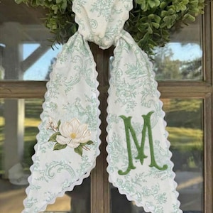 Puede incluir: Un adorno de puerta con estampado floral blanco y verde con una flor de magnolia y la letra "M". El adorno está atado a una corona verde. Los bordes del adorno son festoneados.