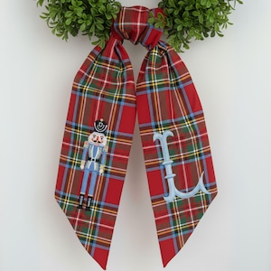 Puede incluir: Bufanda a cuadros rojos con una corona verde. La bufanda presenta un cascanueces bordado y la letra "L" en azul claro. La bufanda está hecha de tela suave, ideal para la temporada navideña.
