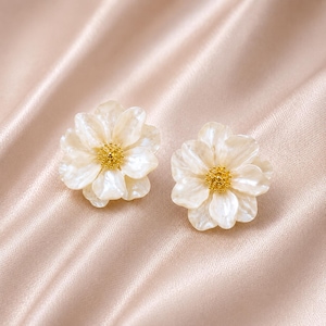 Op de afbeelding: Twee witte bloemvormige oorstekers met een goudkleurig centrum. De oorbellen hebben meerdere bloemblaadjes en een parelmoer afwerking. Ze liggen op een lichtroze satijnen achtergrond.