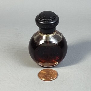 May include: A small, round perfume bottle with a dark brown liquid inside. The bottle has a black cap and sits next to a penny for size comparison. The bottle is made of clear glass.