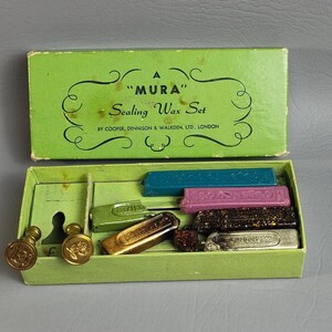 May include: A vintage "MURA" Sealing Wax Set in a green box. The box contains various colored wax sticks, including blue, pink, and gold, along with several brass stampers. The box reads "Sealing Wax Set" and "BY COOPER, DENNISON & WALKDEN, LTD., LONDON."