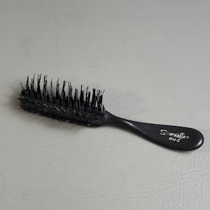 May include: A black hairbrush with a curved handle. The brush has a rectangular head with closely spaced bristles. The handle has the text "Spernette" and Japanese characters. The brush is designed for hair care.