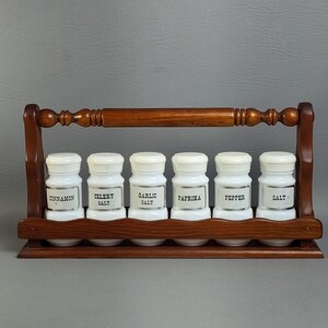 May include: A wooden spice rack with a handle holding six white glass jars. Each jar has a white lid and a label reading Cinnamon, Celery Salt, Garlic Salt, Paprika, Pepper, and Salt. The rack features a dark brown finish and a scalloped base.