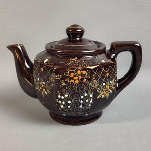 May include: A brown ceramic teapot with a lid and handle. The teapot is decorated with gold accents and floral patterns in orange, white, and blue. The teapot has a spout for pouring and a small base.
