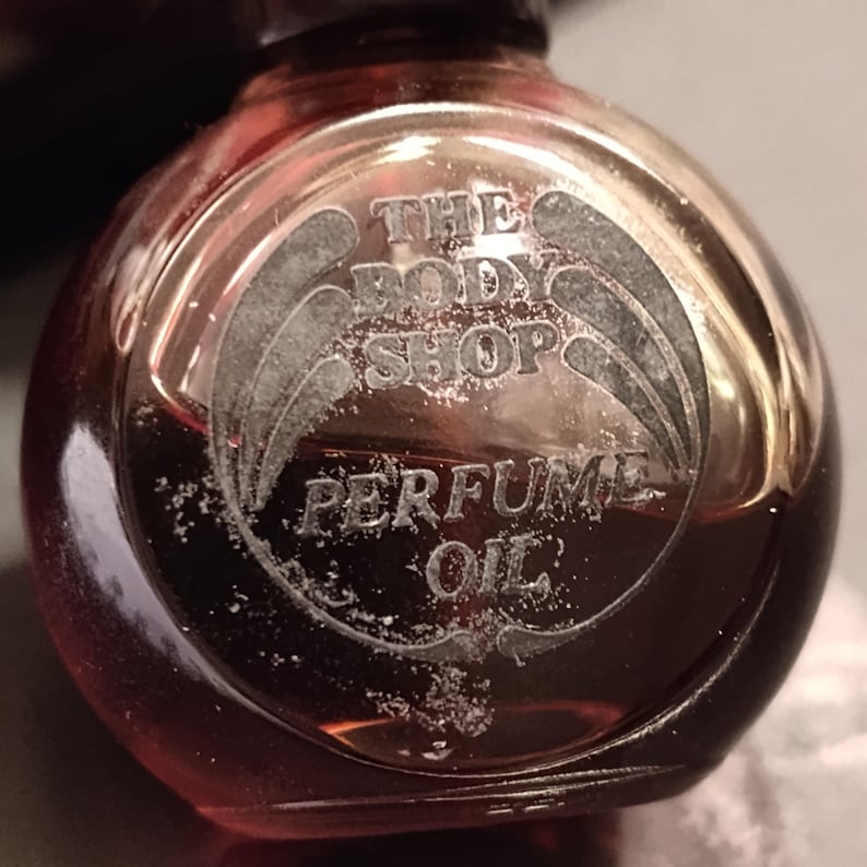 May include: A close-up of a small, round, amber-colored perfume bottle. The bottle has a silver label with the words "THE BODY SHOP PERFUME OIL" etched into it. The bottle is partially filled with a dark liquid.