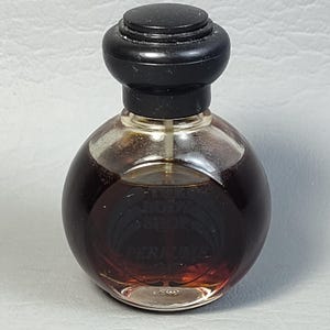 May include: A small, round perfume bottle with a dark brown liquid inside. The bottle has a clear glass body and a black, rounded cap. The label reads "PERFUME". The perfume bottle is a vintage item.