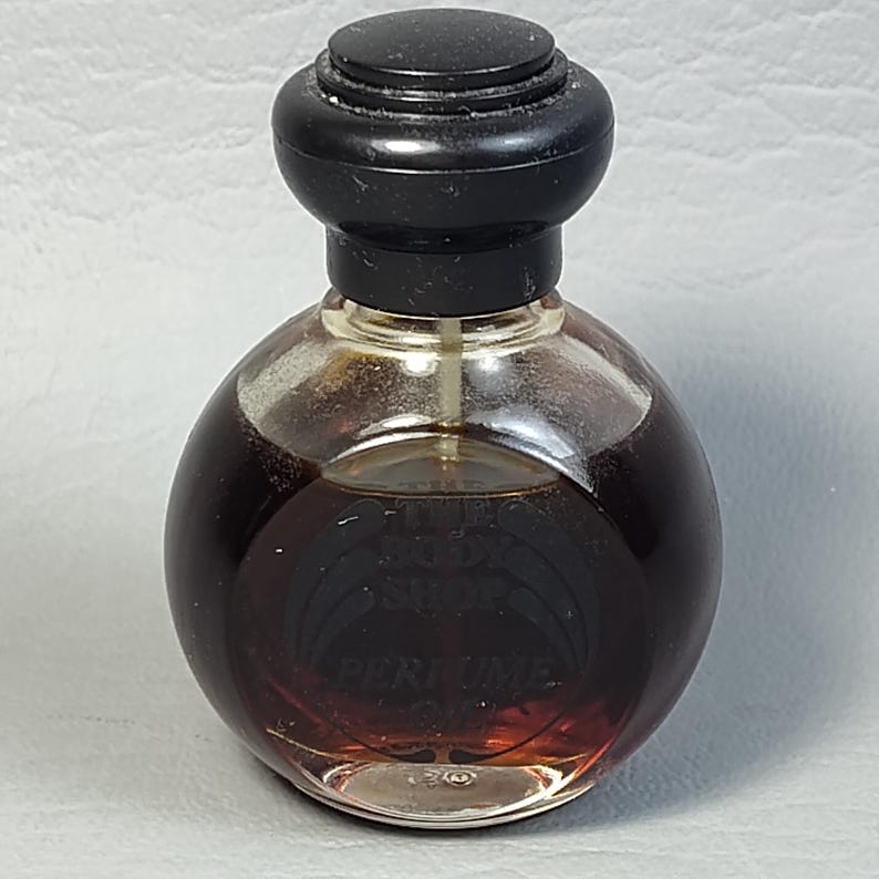 May include: A small, round perfume bottle with a dark brown liquid inside. The bottle has a clear glass body and a black, rounded cap. The label reads "PERFUME". The perfume bottle is a vintage item.