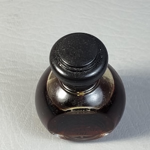 May include: A small, round perfume bottle with a dark brown liquid inside. The bottle has a black, rounded cap. The words "PERFUME OIL" are visible on the bottle. The bottle is sitting on a white surface.