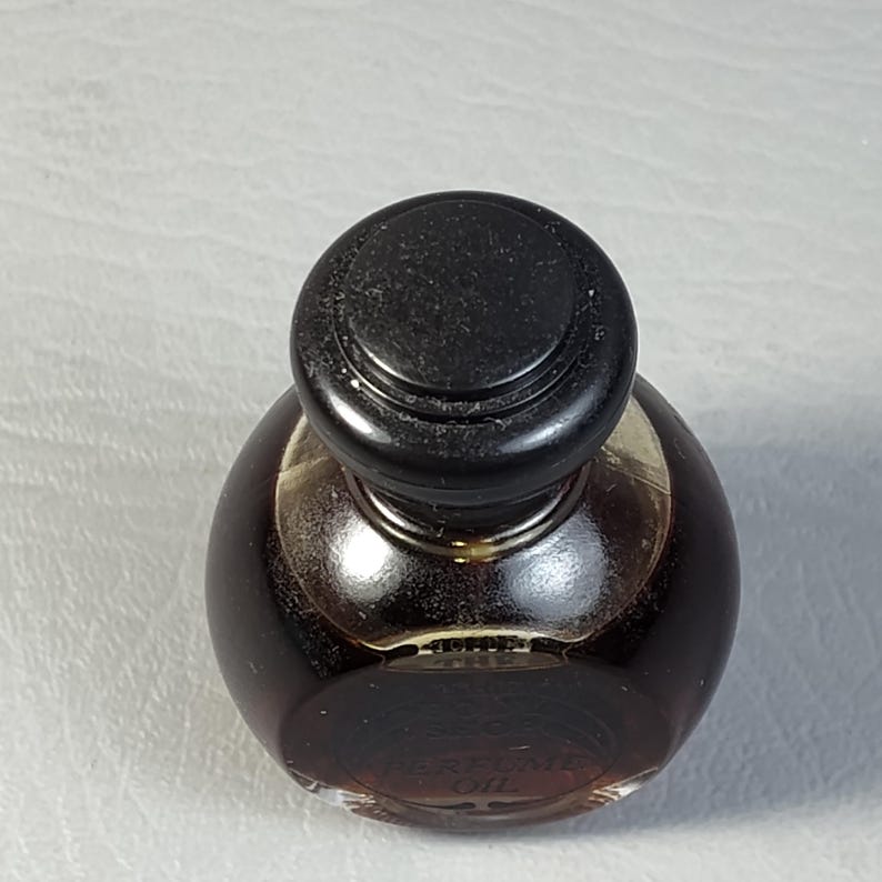 May include: A small, round perfume bottle with a dark brown liquid inside. The bottle has a black, rounded cap. The words "PERFUME OIL" are visible on the bottle. The bottle is sitting on a white surface.