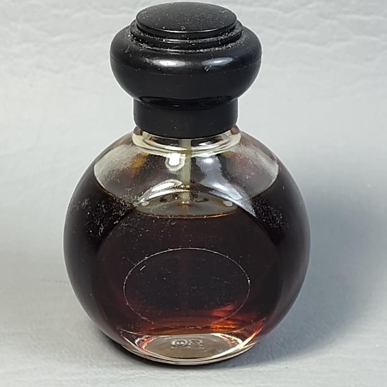 May include: A small, round perfume bottle with a dark brown liquid inside. The bottle has a clear glass body and a black, rounded cap. The perfume bottle is likely vintage, with a classic design.