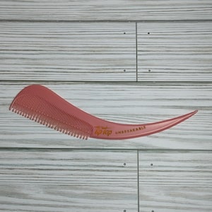 May include: A pink comb with a curved handle and fine teeth. The comb has the words "Tip-Top UNBREAKABLE" printed in gold lettering. The comb is made of a shiny, smooth plastic material.