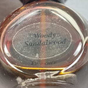 May include: A clear, round perfume bottle with a dark brown cap. The bottle has a label that reads "Woody Sandalwood". The liquid inside is amber-colored. The bottle is partially in focus, with the label being the clearest part.