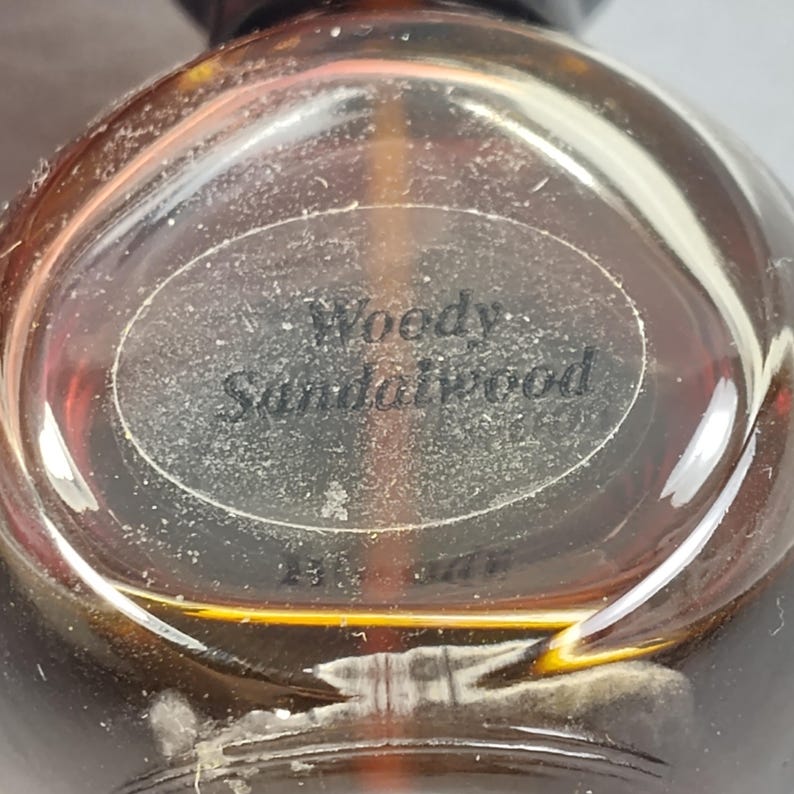 May include: A clear, round perfume bottle with a dark brown cap. The bottle has a label that reads "Woody Sandalwood". The liquid inside is amber-colored. The bottle is partially in focus, with the label being the clearest part.