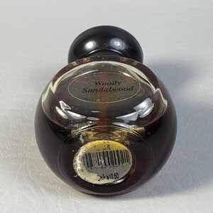 May include: A small, round perfume bottle with a dark brown glass body and a black, spherical cap. The label reads "Woody Sandalwood." A barcode sticker is affixed to the bottom.