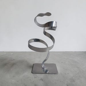May include: A modern, abstract sculpture made of polished metal. The sculpture features a swirling, ribbon-like design that rises from a rectangular base. The metal has a reflective, silver finish, creating a sleek and contemporary aesthetic.