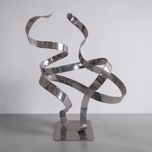 May include: A polished silver-toned metal sculpture featuring a series of interconnected, flowing ribbons that twist and curve upward from a flat, square base. The reflective surface creates smooth, continuous lines that form an abstract shape.