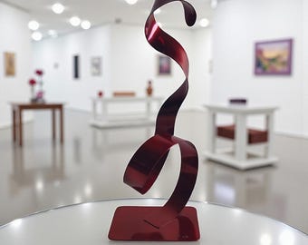 Op de afbeelding: Een moderne, abstracte sculptuur van glanzend, rood metaal. De sculptuur heeft een rechthoekige basis en een lintachtige vorm die omhoog buigt en draait. Het kunstwerk wordt tentoongesteld op een witte tafel in een galerie.