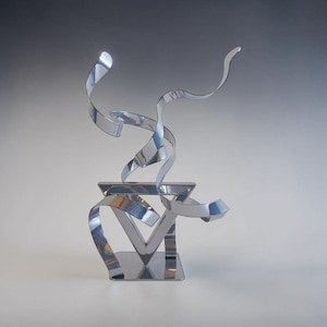 Op de afbeelding: Een sculptuur van gepolijst zilverkleurig metaal met een platte vierkante voet die een centraal driehoekig frame ondersteunt. Verschillende gebogen, lintachtige metalen strips steken vanuit het midden omhoog en naar buiten, waardoor een dynamische, multidimensionale vorm ontstaat met reflecterende oppervlakken over de gehele structuur.
