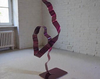 Op de afbeelding: Een moderne, abstracte sculptuur in een levendige magenta tint. Het kunstwerk toont een doorlopende, lintachtige vorm die zich omhoog draait en buigt vanaf een rechthoekige basis. Het gepolijste metaal reflecteert licht, waardoor een dynamisch visueel effect ontstaat. Geschikt voor interieurdecoratie.