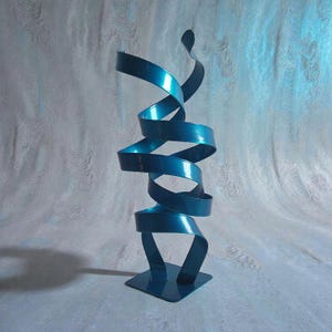 Pode incluir: Uma escultura de metal azul-petróleo com um design de fita em espiral. A escultura tem uma base quadrada e uma forma contínua e fluida que se torce para cima. A cor azul-petróleo tem um acabamento brilhante, refletindo a luz. A obra de arte é uma peça moderna e abstrata.