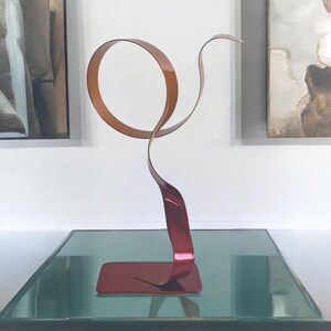 Può includere: Una scultura astratta moderna nei toni del rosso e del rame. L'opera d'arte in metallo presenta una base rettangolare, uno stelo curvo e un anello circolare nella parte superiore. La scultura è esposta su una superficie di vetro.